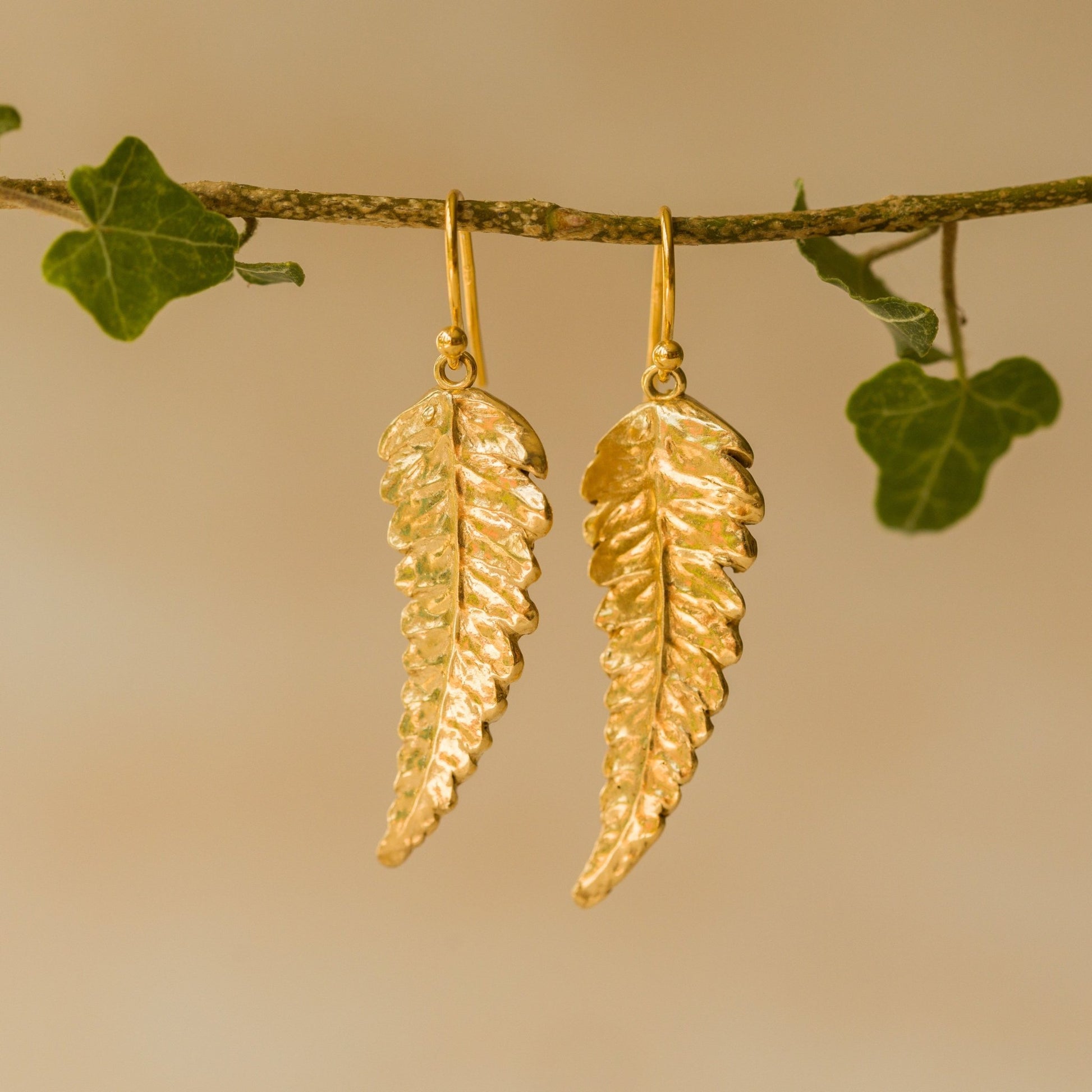 Handmade recycled gold Fern Leaf earrings by Iris and Lolli hanging from an ivy branch, featuring intricate fern detail and a radiant golden woodland glow