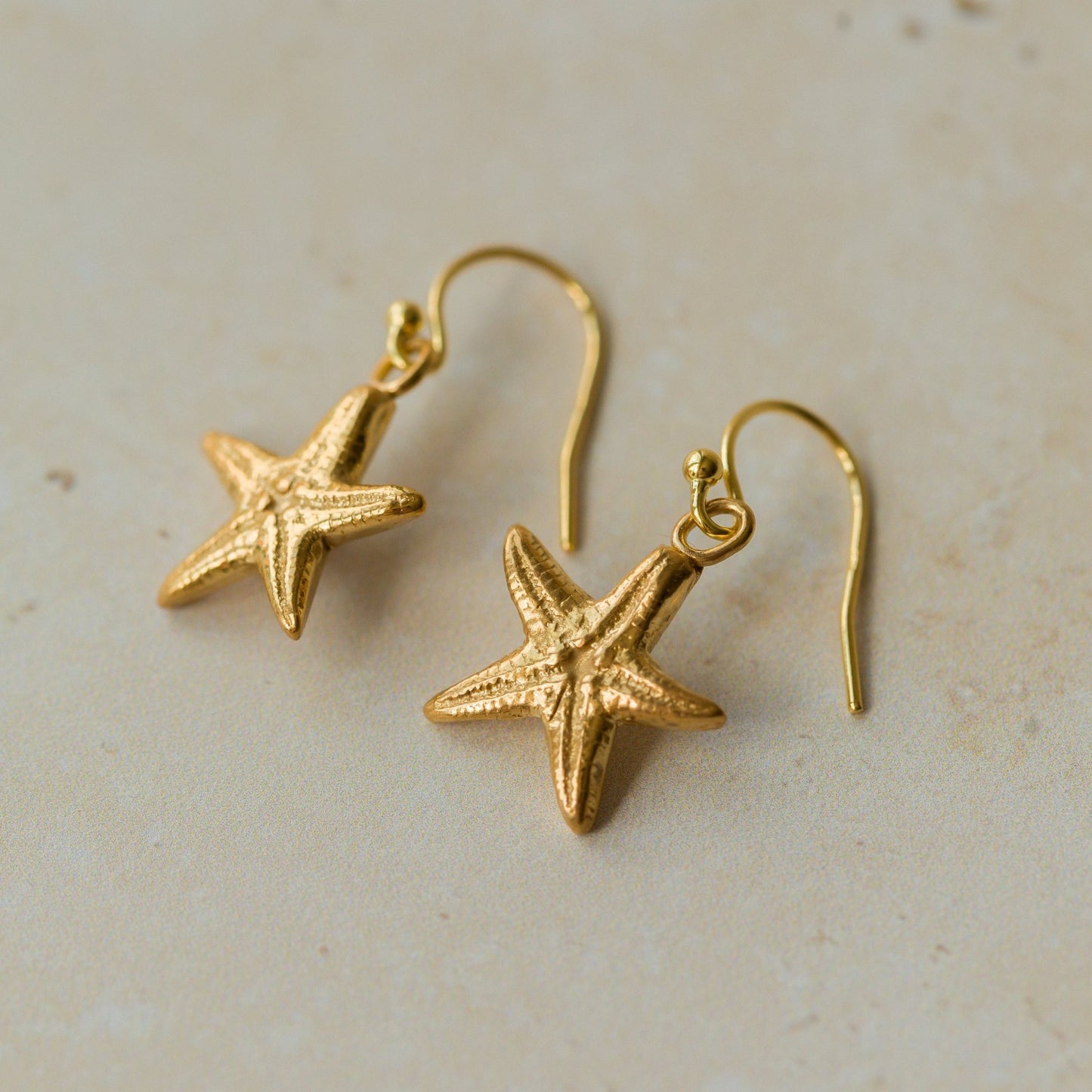 Handmade recycled gold Starfish earrings by Iris and Lolli laid flat on a sandy-toned background, showing textured sea star detail and coastal-inspired shine