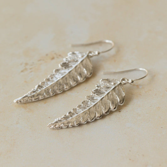 Close-up of handmade recycled silver Fern earrings by Iris and Lolli on a neutral stone background, showing delicate leaf texture and natural botanical detail