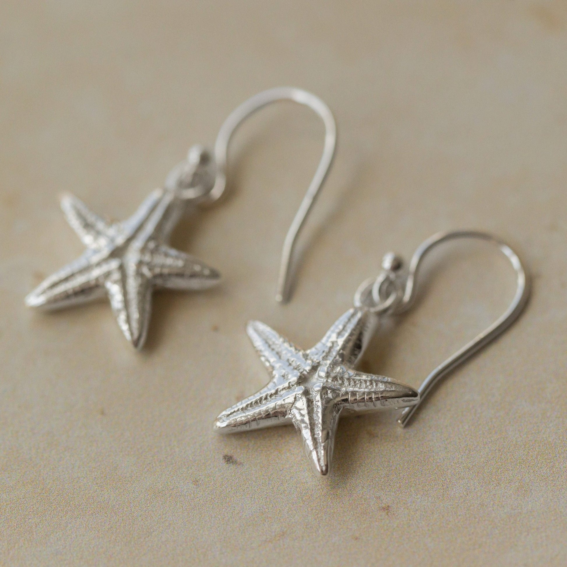 Close-up of handmade recycled silver Starfish earrings by Iris and Lolli resting on a neutral stone surface, showing textured sea-inspired design and delicate hooks