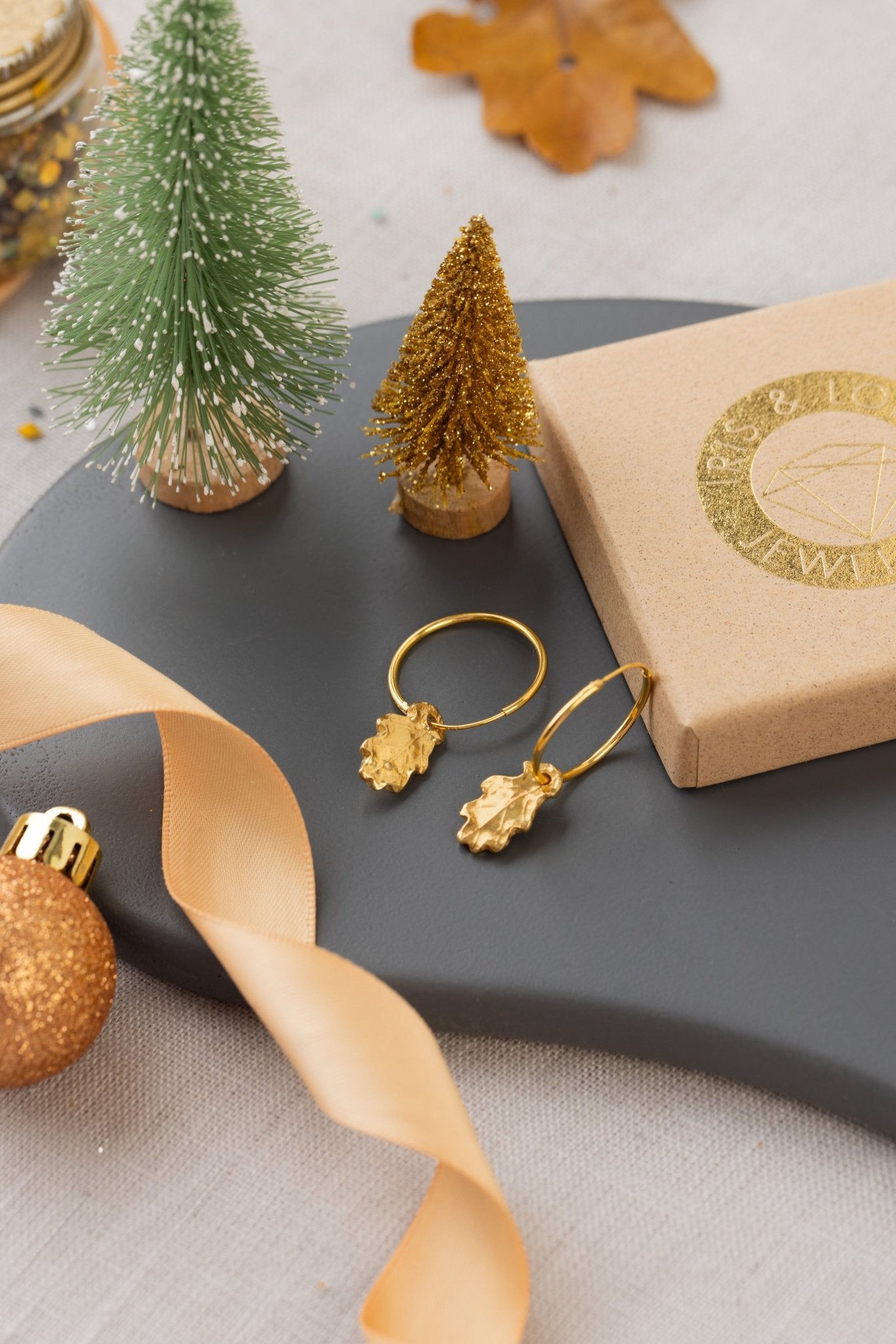 Gold earrings on a dark surface with decorative items including a small Christmas tree and ribbon.