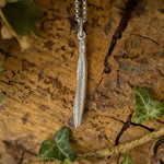 Silver leaf pendant necklace on a wooden surface with green leaves.