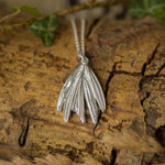Silver leaf-shaped pendant on a chain, placed on a wooden surface with green leaves and dry grass.