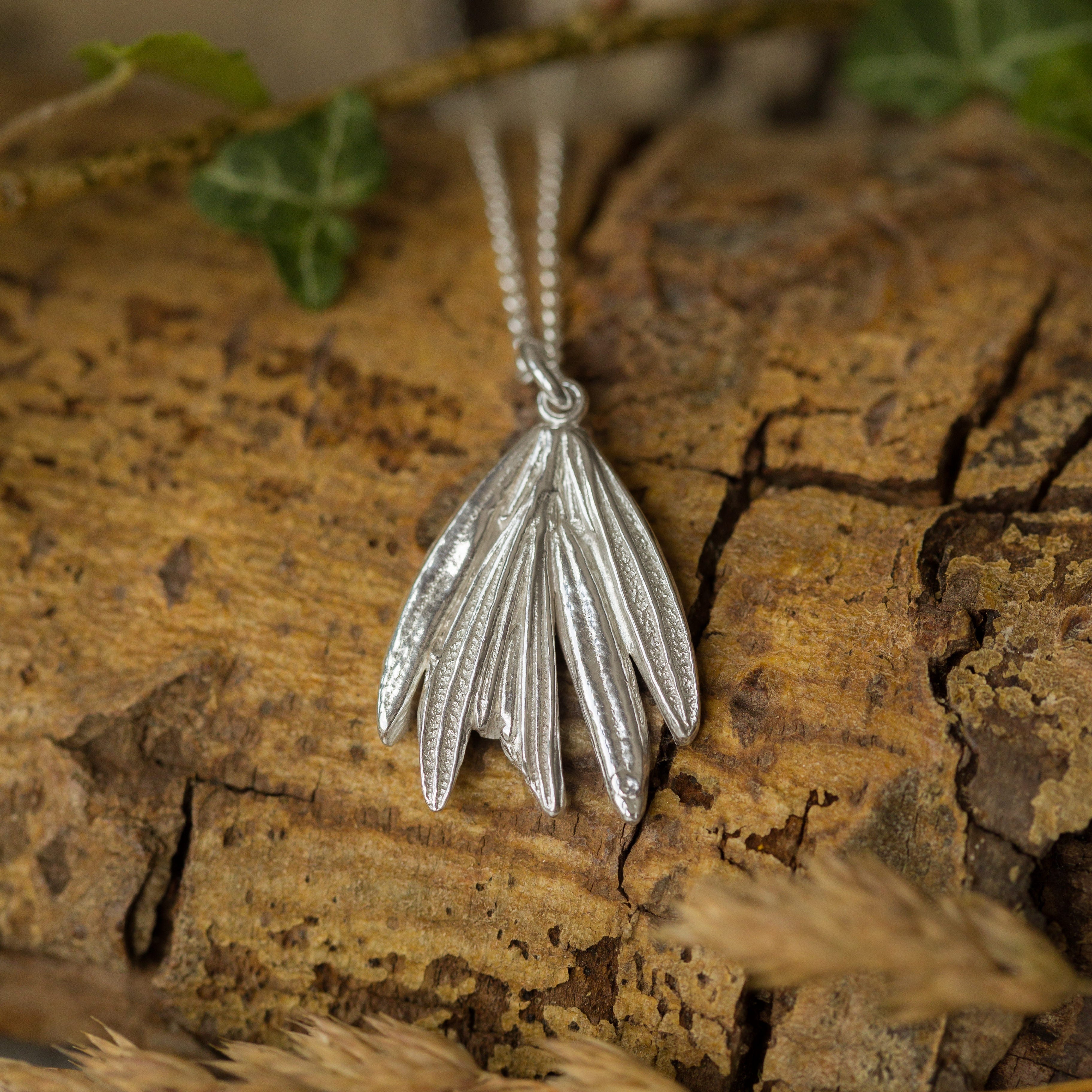 Silver leaf-shaped pendant on a chain, placed on a wooden surface with green leaves and dry grass.