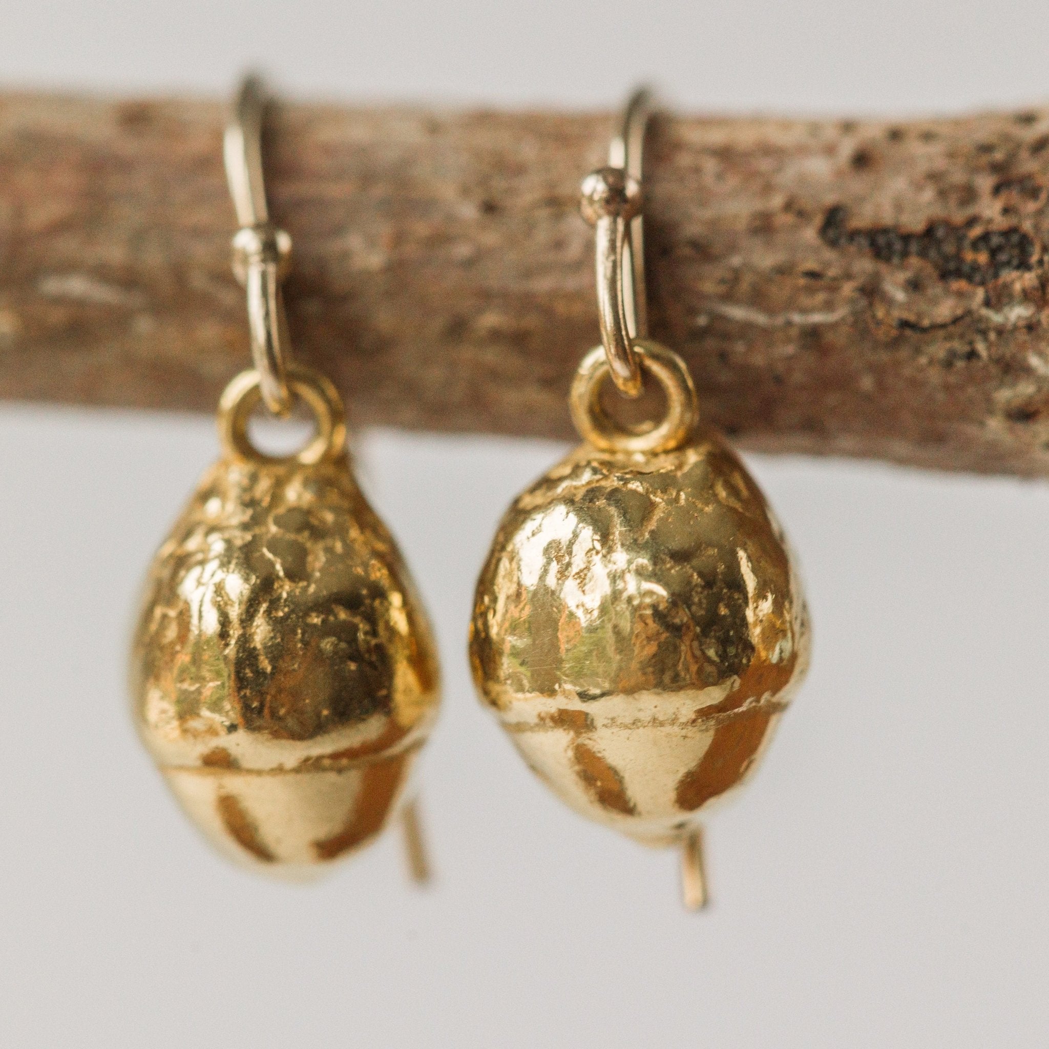 close up of gold acorn drop earrings with textured finish, handcrafted nature inspired jewellery displayed on wooden twig