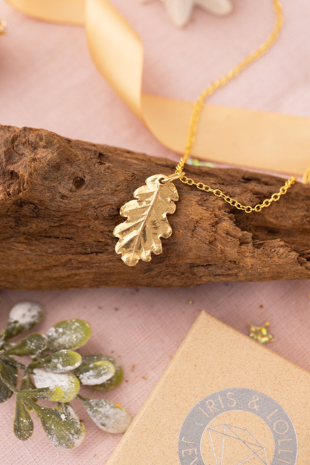 Gold oak leaf necklace resting on driftwood with a gold ribbon and Christmas decorations