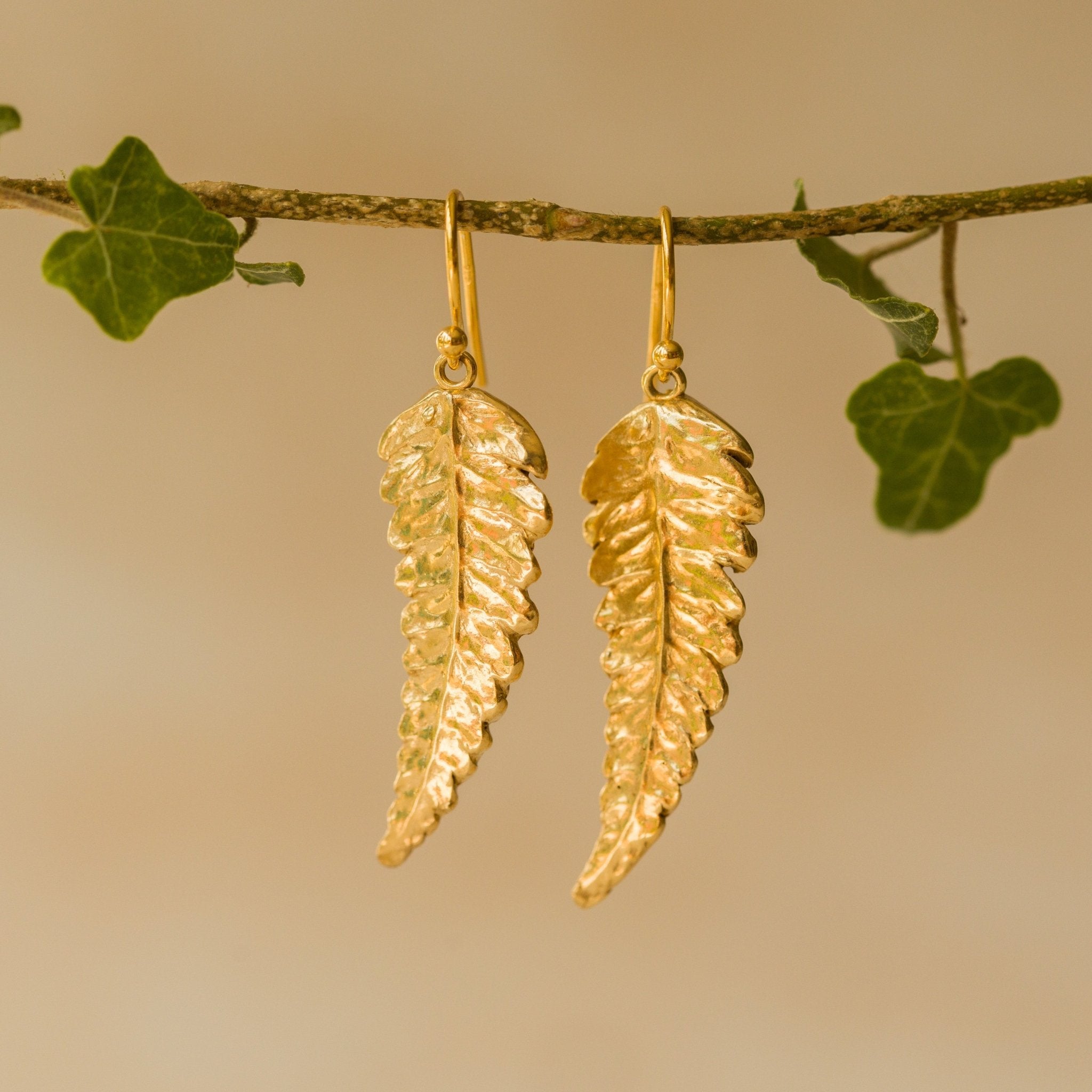 Handmade recycled gold Fern Leaf earrings by Iris and Lolli hanging from an ivy branch, featuring intricate fern detail and a radiant golden woodland glow