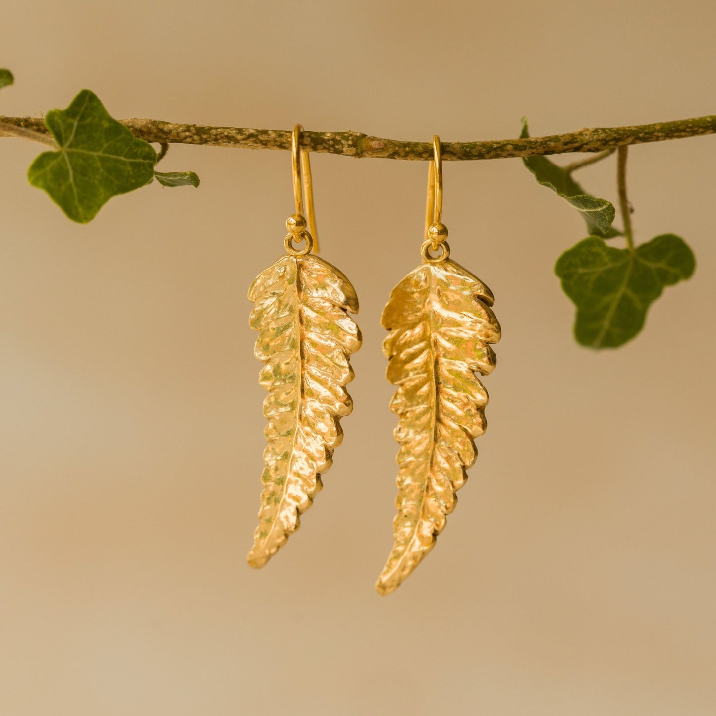 Handmade recycled gold Fern Leaf earrings by Iris and Lolli hanging from an ivy branch, featuring intricate fern detail and a radiant golden woodland glow