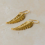 Close-up of handmade recycled gold Fern Leaf earrings by Iris and Lolli on a neutral stone background, showing delicate leaf texture and natural curved detailing