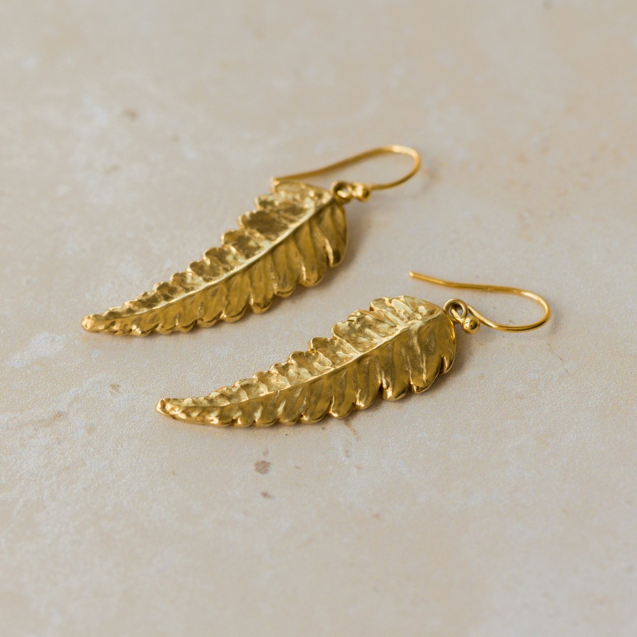 Close-up of handmade recycled gold Fern Leaf earrings by Iris and Lolli on a neutral stone background, showing delicate leaf texture and natural curved detailing