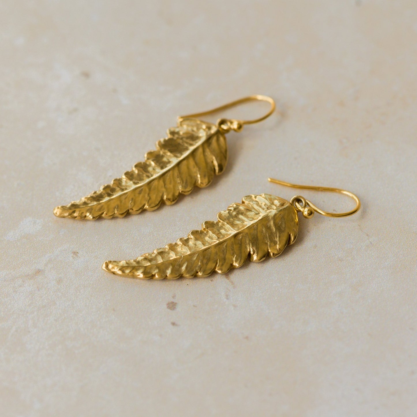 Close-up of handmade recycled gold Fern Leaf earrings by Iris and Lolli on a neutral stone background, showing delicate leaf texture and natural curved detailing
