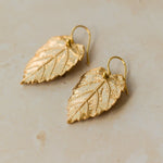 Close-up of handmade recycled gold Hazel Leaf earrings by Iris and Lolli on a soft neutral surface, showing intricate leaf veins and a rich golden finish