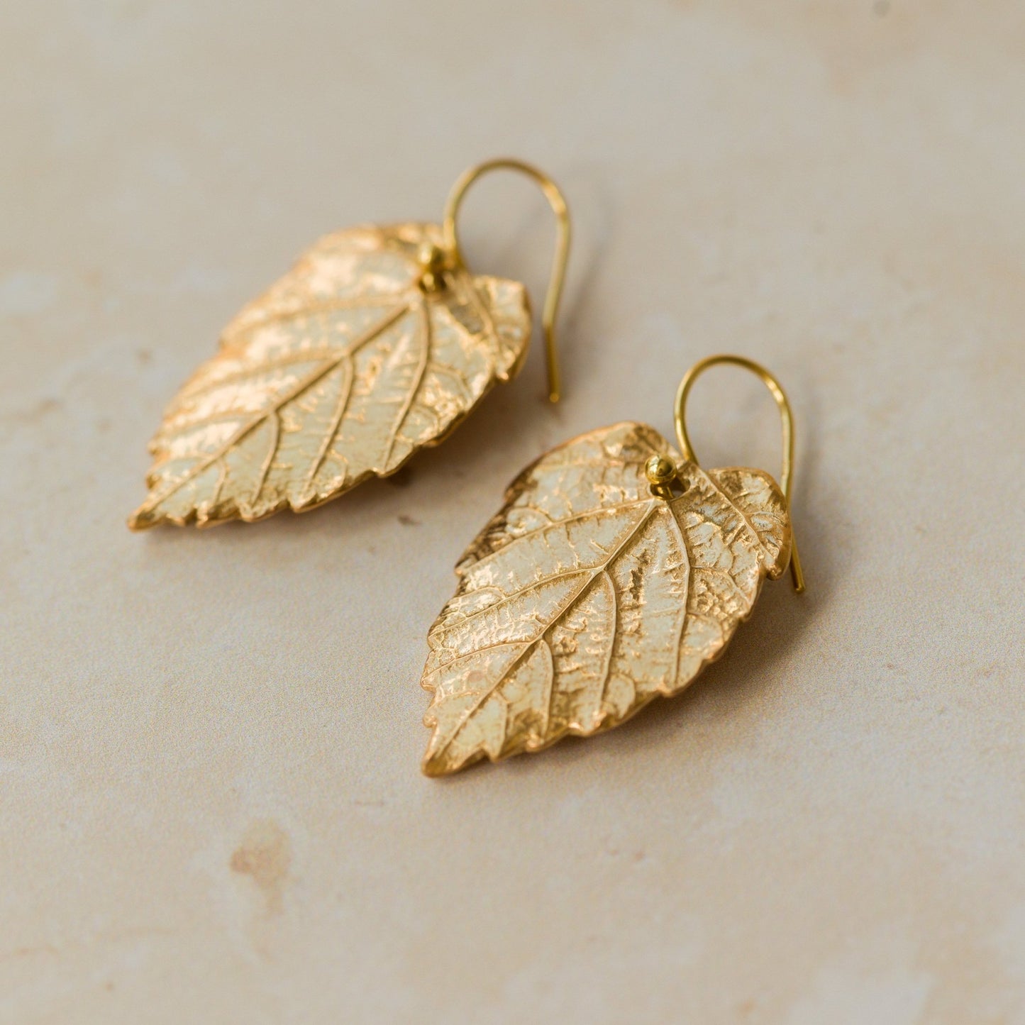 Close-up of handmade recycled gold Hazel Leaf earrings by Iris and Lolli on a soft neutral surface, showing intricate leaf veins and a rich golden finish