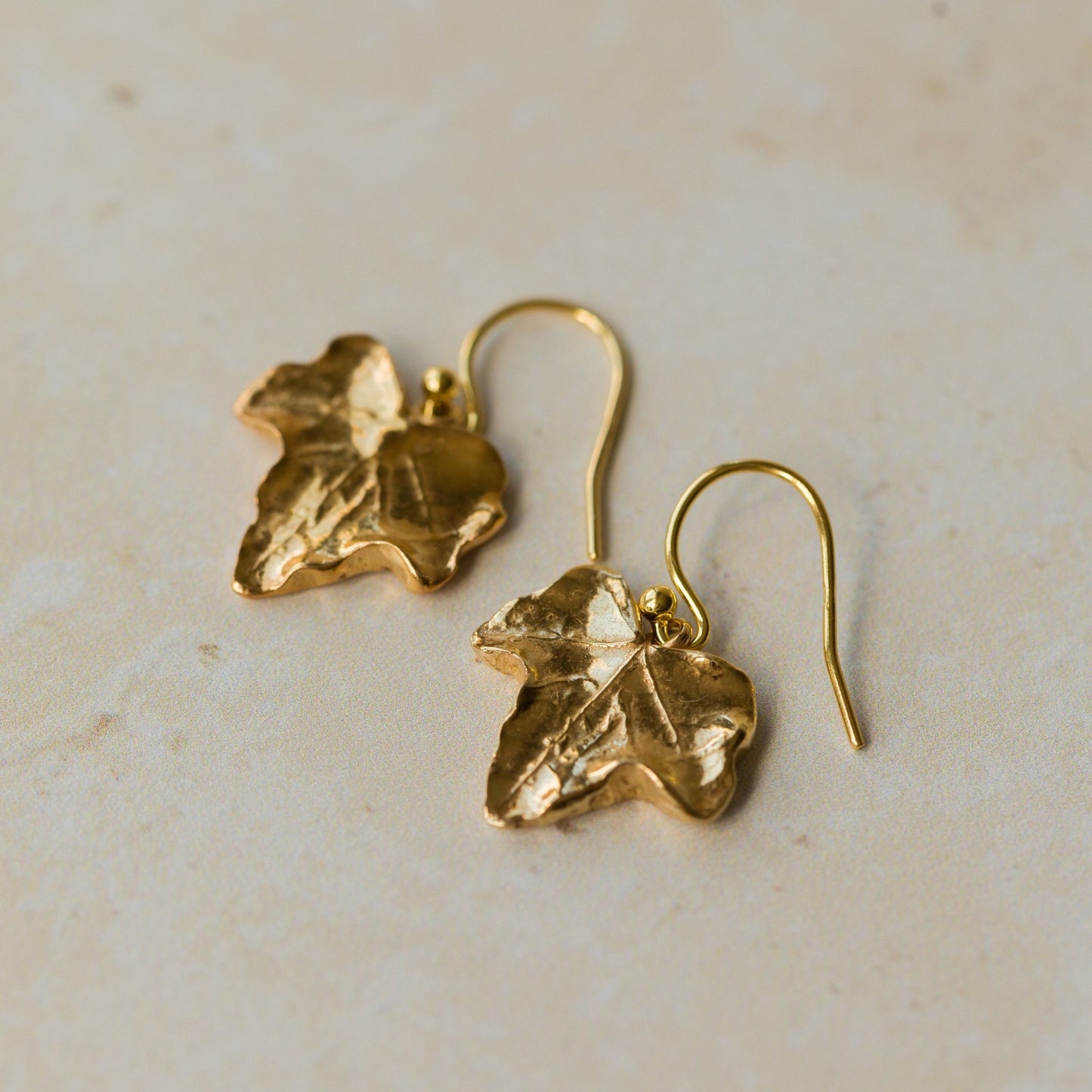 Close-up of handmade recycled gold Ivy Leaf earrings by Iris and Lolli on a warm neutral background, showing their natural leaf texture and soft golden sheen