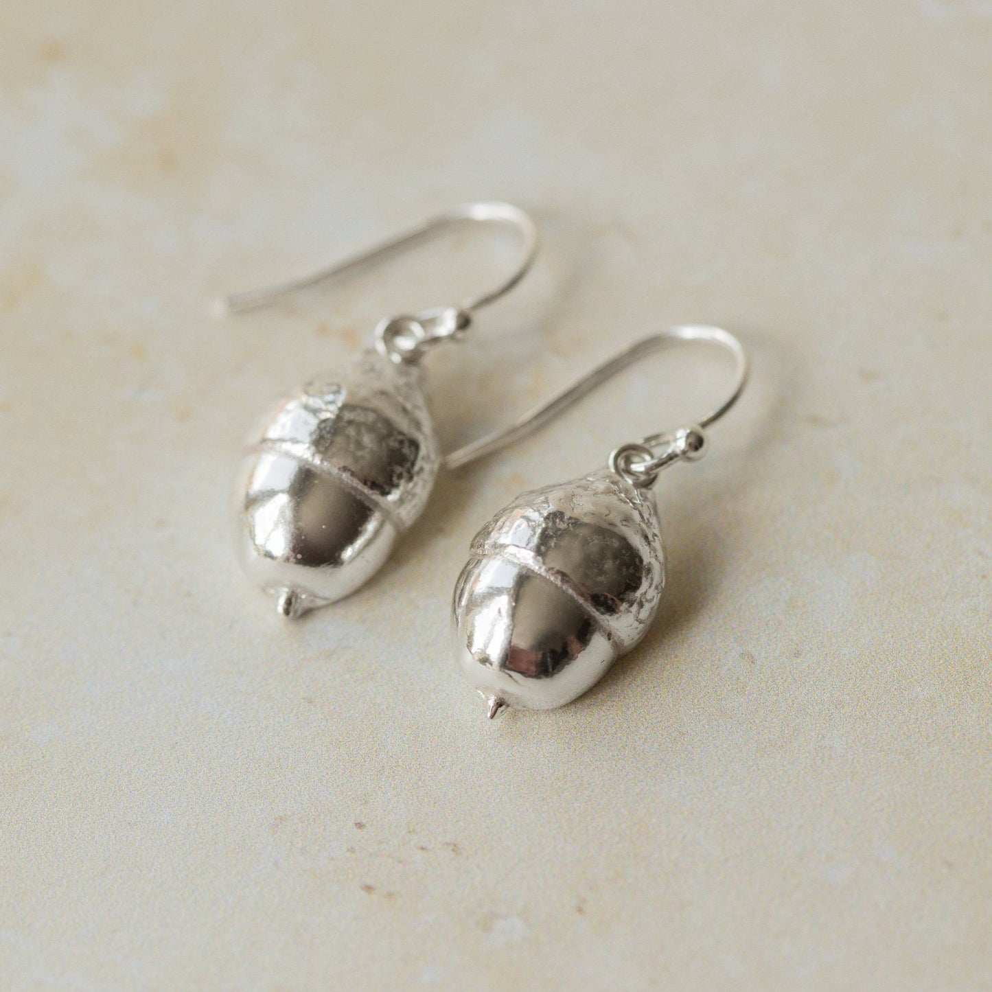 Close-up of handmade recycled silver Acorn earrings by Iris and Lolli on a neutral surface, highlighting the detailed acorn texture and polished silver finish