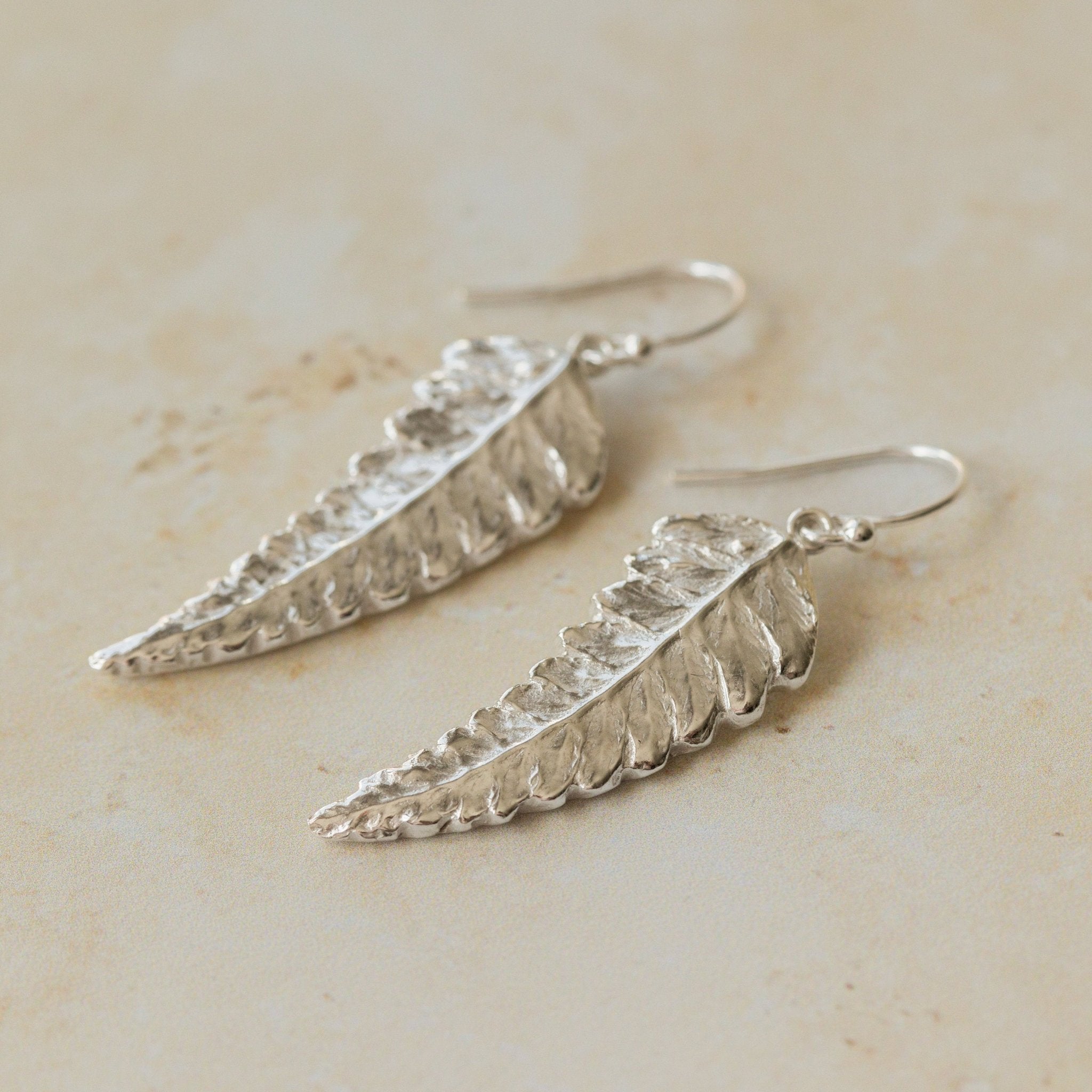 Close-up of handmade recycled silver Fern earrings by Iris and Lolli on a neutral stone background, showing delicate leaf texture and natural botanical detail