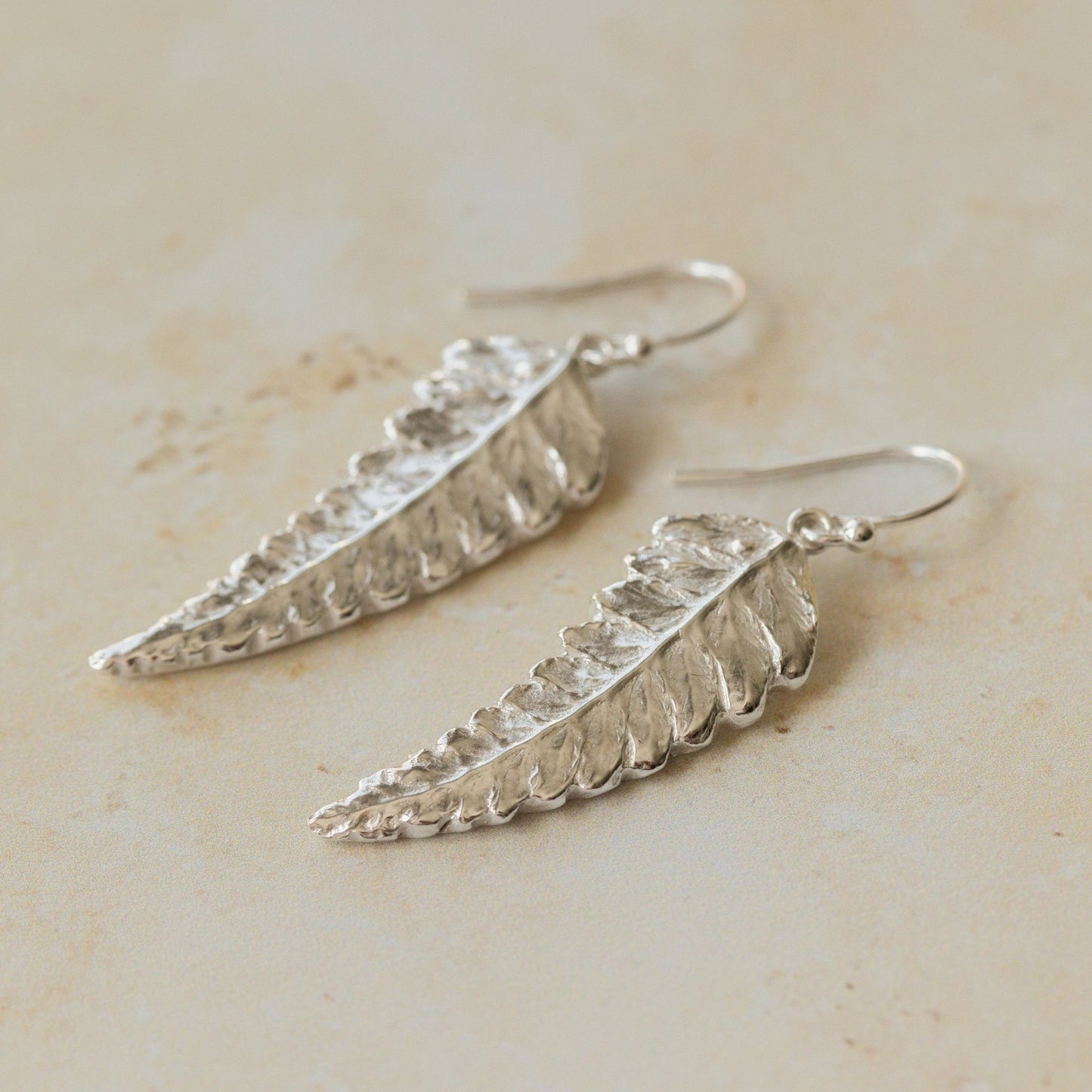Close-up of handmade recycled silver Fern earrings by Iris and Lolli on a neutral stone background, showing delicate leaf texture and natural botanical detail