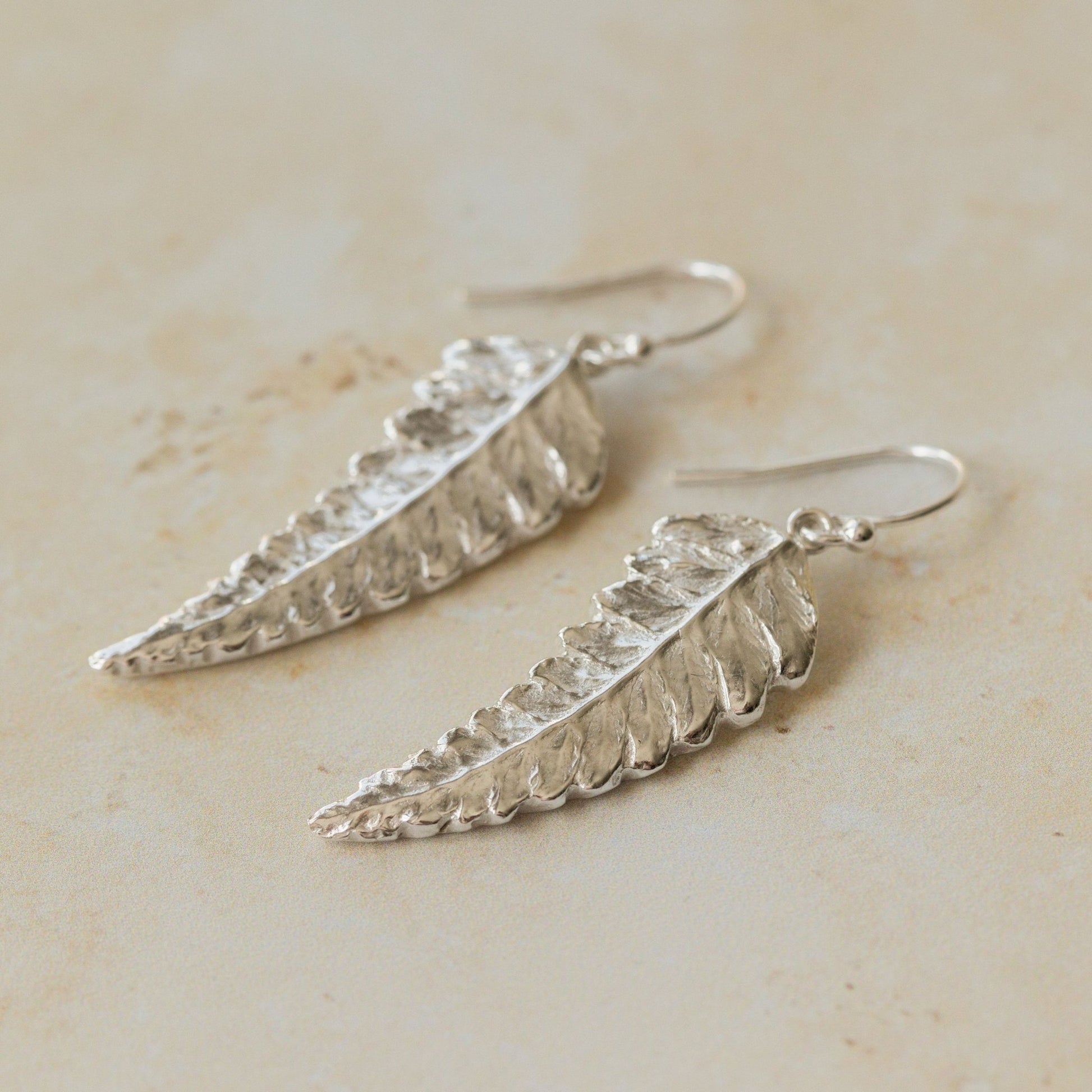 Close-up of handmade recycled silver Fern earrings by Iris and Lolli on a neutral stone background, showing delicate leaf texture and natural botanical detail