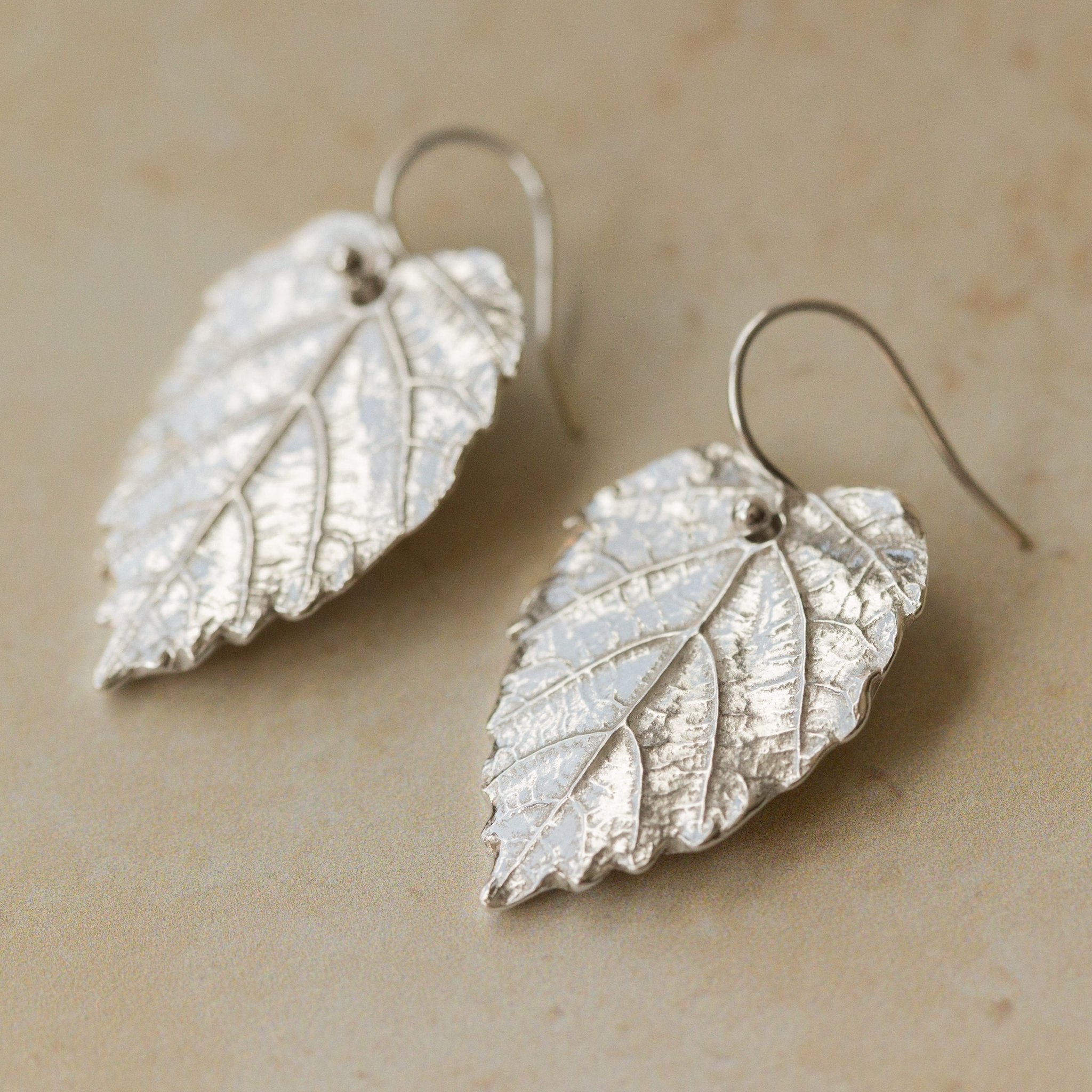 Close-up of handmade recycled silver Hazel Leaf earrings by Iris and Lolli on a neutral stone surface, showing detailed leaf veins and organic texture that capture the natural shape of the hazel leaf