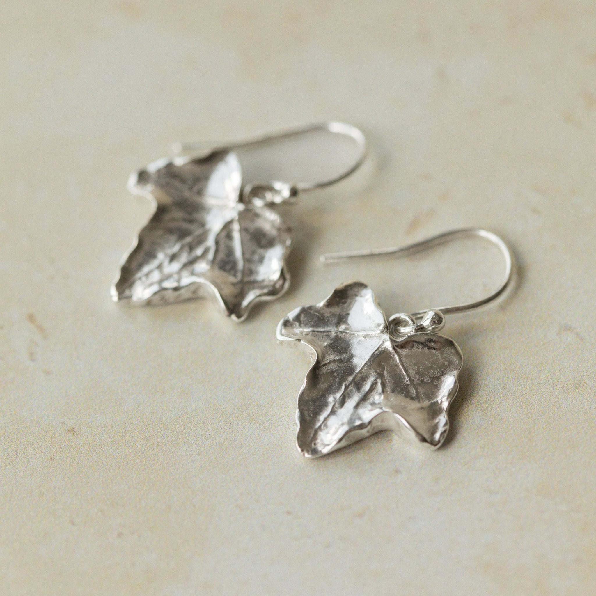 Close-up of handmade recycled silver Ivy Leaf earrings by Iris and Lolli on a soft neutral surface, highlighting the natural leaf texture and organic botanical shape