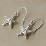 Close-up of handmade recycled silver Starfish earrings by Iris and Lolli resting on a neutral stone surface, showing textured sea-inspired design and delicate hooks