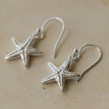 Close-up of handmade recycled silver Starfish earrings by Iris and Lolli resting on a neutral stone surface, showing textured sea-inspired design and delicate hooks