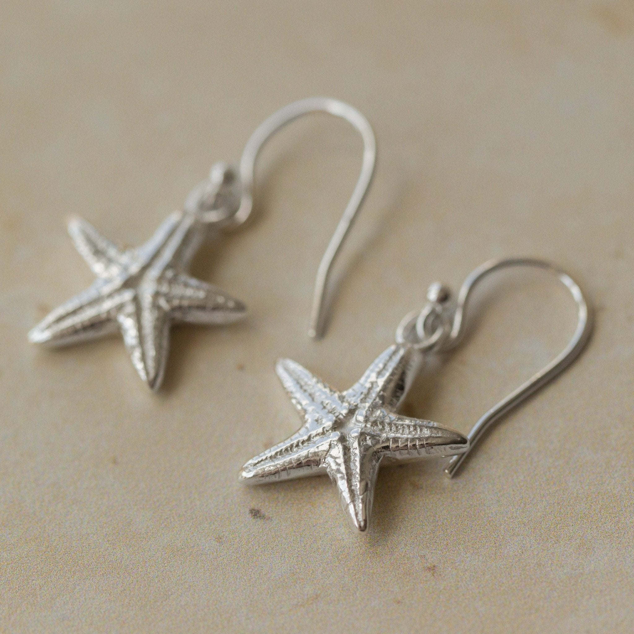 Close-up of handmade recycled silver Starfish earrings by Iris and Lolli resting on a neutral stone surface, showing textured sea-inspired design and delicate hooks