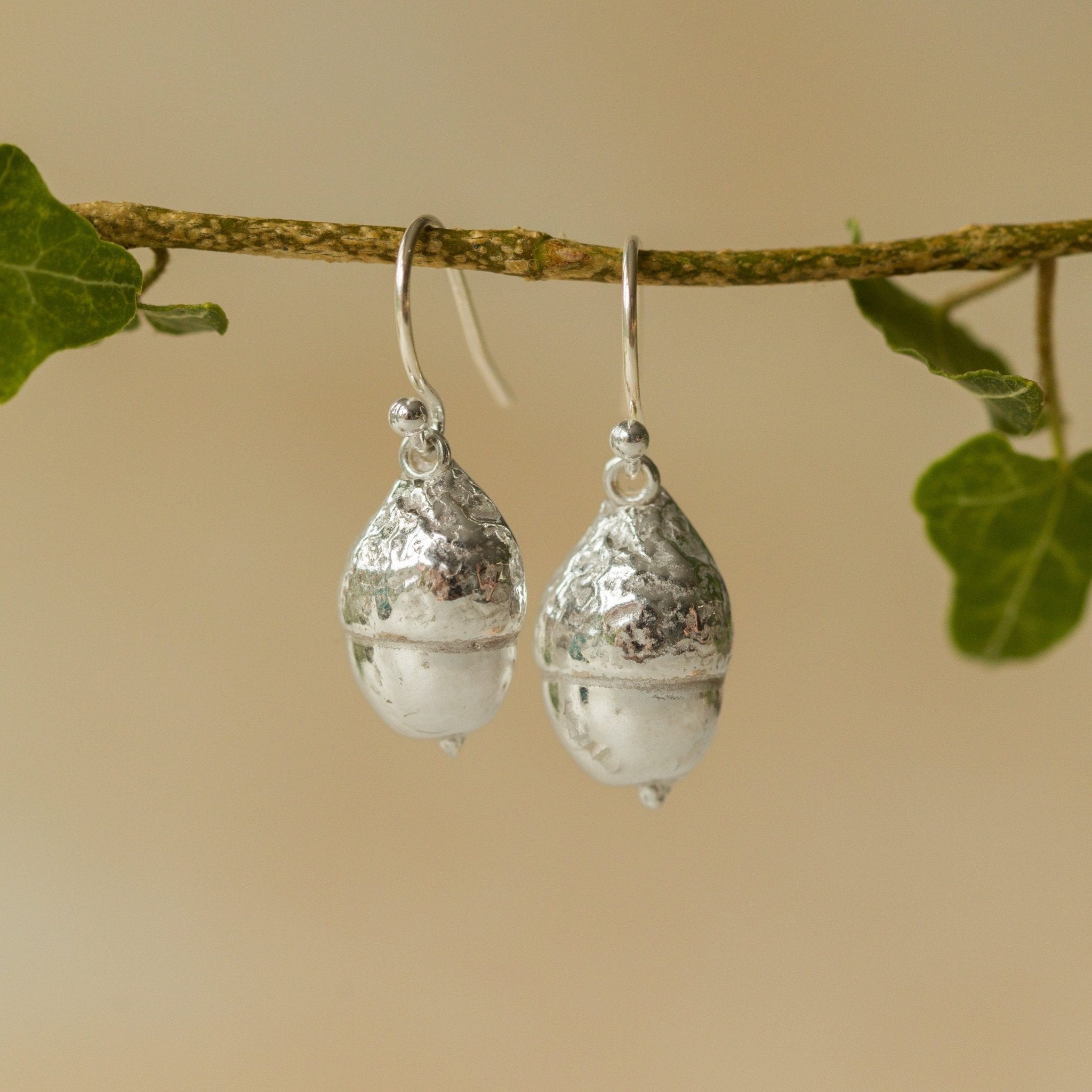 Handmade recycled silver Acorn earrings by Iris and Lolli hanging from a branch with ivy leaves, showcasing their natural woodland texture and polished silver shine