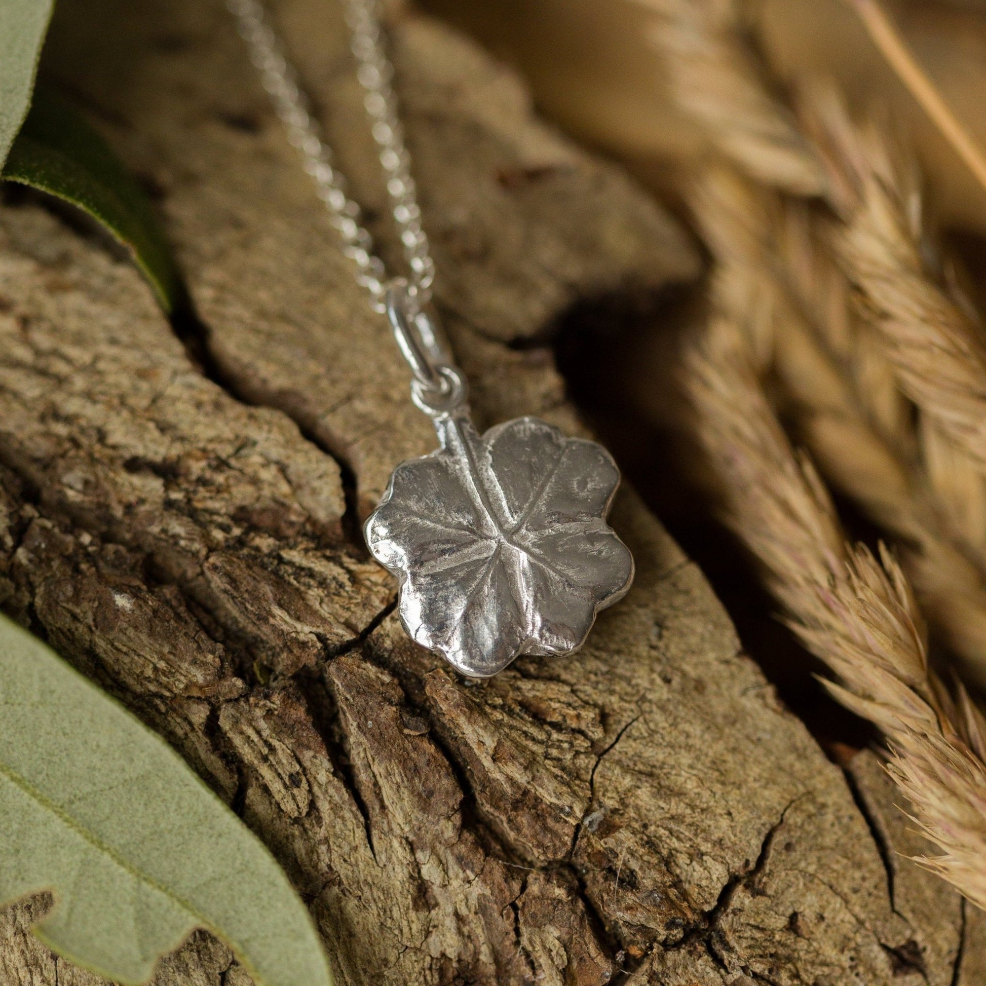 Handcrafted recycled-silver clover leaf necklace displayed on natural bark. Textured botanical pendant symbolising luck and nature, a meaningful jewellery gift.