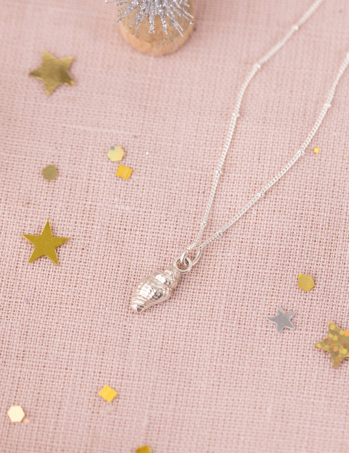 Silver dainty whelk shell necklace on pink linen with gold stars